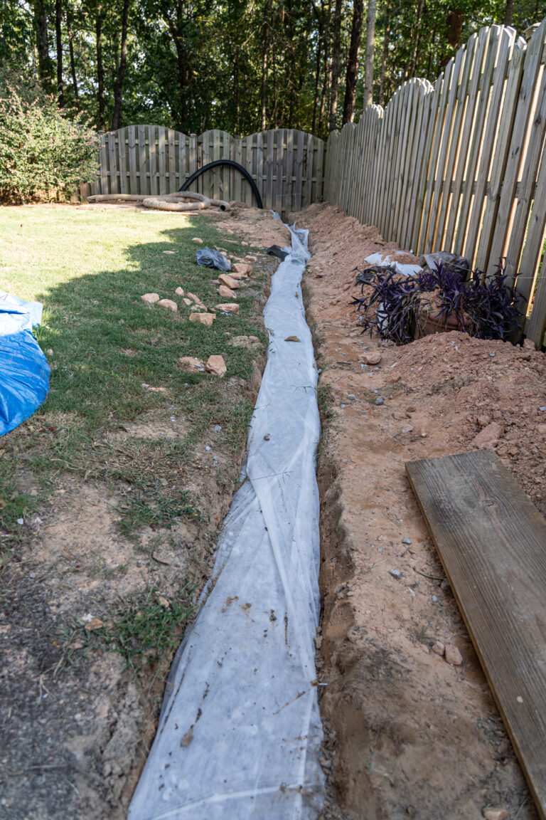 Fabric covers drainage rock and pipe to keep a French drain clear of debris in this DIY home improvement project to alleviate drainage issues.